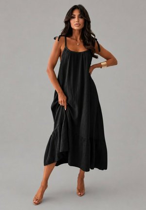 Femme aux longs cheveux foncés portant une robe maxi noire sans manches avec des bretelles à nouer, posant pieds nus devant un fond gris uni.