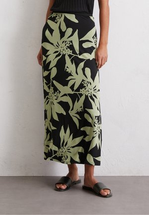 Kobieta nosząca czarną spódnicę midi z dużymi zielonymi kwiatowymi wzorami, w połączeniu z czarnymi sandałami typu slide, stoi na tle białej ściany.
