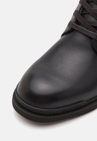 Chaussure en cuir noir avec une finition lisse, bout rond, design à lacets foncés et semelle en caoutchouc présentant un accent de marque sur le côté.