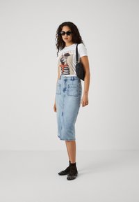Fustă tip creion din denim albastru, cu două buzunare frontale, asortată cu un tricou alb cu grafică și balerini negri cu ținte. Ochelari de soare negri și o geantă mică.