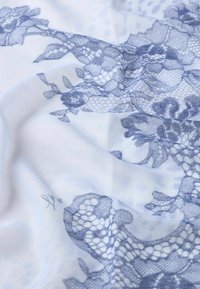 Tessuto leggero con sfondo bianco caratterizzato da intricate fantasie di pizzo blu. Presenta una texture morbida con delicati accenti floreali.