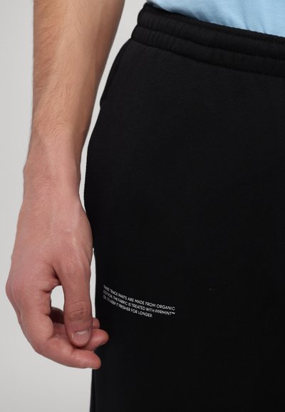 Black track pants made from organic cotton, featuring an elastic waistband and a white text label on the side with care information.