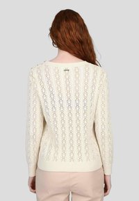 Pull en tricot de couleur crème avec un motif en dentelle, manches longues et poignets côtelés. La vue de dos met en avant un détail de boutons près de l'épaule.