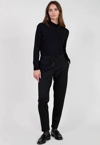 Zwarte top met lange mouwen en een col hals, gecombineerd met zwarte taps toelopende broek met elastische tailleband en zijzakken. Draagt zwarte schoenen.