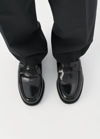 Μαύρο δερμάτινο λόφο με λεία υφή, με πλεγμένες λωρίδες και στρογγυλή μύτη, συνδυασμένο με φαρδιά μαύρα παντελόνια.