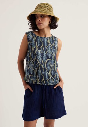 Vrouw die een mouwloze marineblauwe top draagt met groen-blauwe bladpatronen, marineblauwe textuurshort, een groene en beige geweven bucket hat, en gouden oorringen.