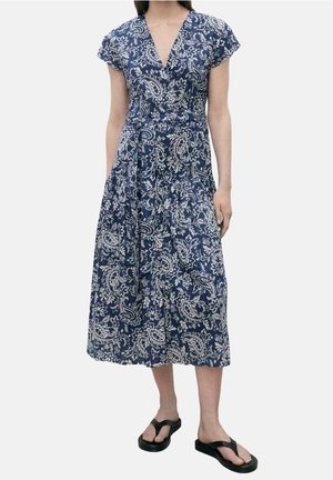 Femme portant une robe midi bleue avec des motifs paisley blancs et des manches courtes, associée à des sandales noires ouvertes au niveau des orteils.