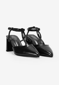 Tacones de charol negros, de punta, con tirante al tobillo y tacones de bloque. Presentan un diseño con lados abiertos para un detalle adicional.