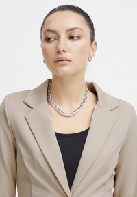 Blazer beige sur un top noir, avec un collier en chaîne torsadée argenté mettant en valeur le décolleté. Tissu lisse et design structuré.