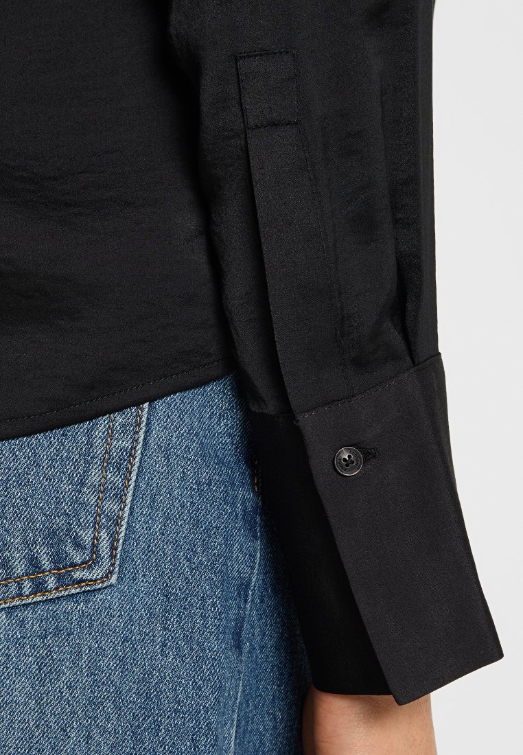 Zwarte shirt met een gladde textuur, met een taps toelopende mouw en een enkele knoopsluiting bij de manchet, gecombineerd met denim jeans.