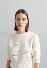Weißer Strickpullover mit einem strukturierten Design und kurzen Ärmeln, der horizontale und vertikale Muster sowie einen weiten Ausschnitt aufweist.