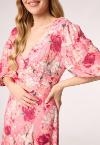 Robe florale composée d'un tissu rose avec des fleurs rouges et blanches, un décolleté en V profond, des manches bouffantes remarquables et une taille cintrée.