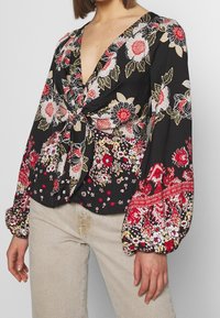 Black floral blouse with a deep V-neck, knot detail at the waist, and long puffed sleeves featuring red, white, and yellow patterns.