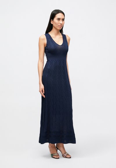 Tmavomodré maxi šaty bez rukávov s textúrovaným geometrickým vzorom, priliehavým živôtikom a rozšírenou sukňou, doplnené viacfarebnými sandálmi na podpätku so remienkami.