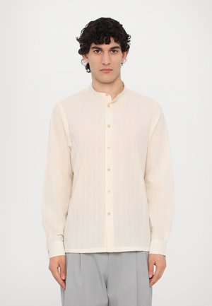 Camisa de manga larga beige claro con cuello de banda, con cierre de botones y tela sutilmente texturizada. Combinada con pantalones grises.