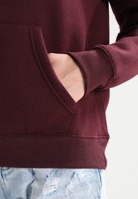 Sudadera marrón con bolsillo canguro, puños y cintura acanalados. Tejido texturizado, combinada con jeans de mezclilla azul claro desgastados.
