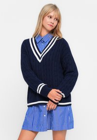 Marineblauer Zopfstrickpullover mit V-Ausschnitt, weißen Streifenakzenten am Ausschnitt und den Bündchen, kombiniert mit einem blauen gestreiften Hemd und einem Rock.
