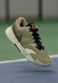 Nike sneakers ar bēša un melnā krāsu shēmu, teksturētu mežģīņu augšdaļu, apaļu purngalu, gumijas zoli un logotipa uzlīmi uz mēles.