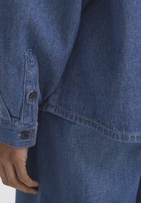 Chemise en denim avec poignets à boutons, de couleur bleue, coutures claires et coupe décontractée. Le tissu a une texture lisse.