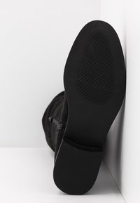 Botte noire à semelle plate reposant sur le côté, montrant la semelle texturée et la fermeture éclair latérale sur un fond blanc en coin.