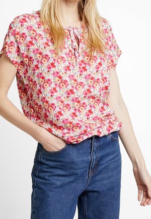 Blusa floral en rosa, naranja y amarillo con mangas cortas, cuello redondo y un detalle de lazo en el cuello, combinada con jeans de mezclilla.