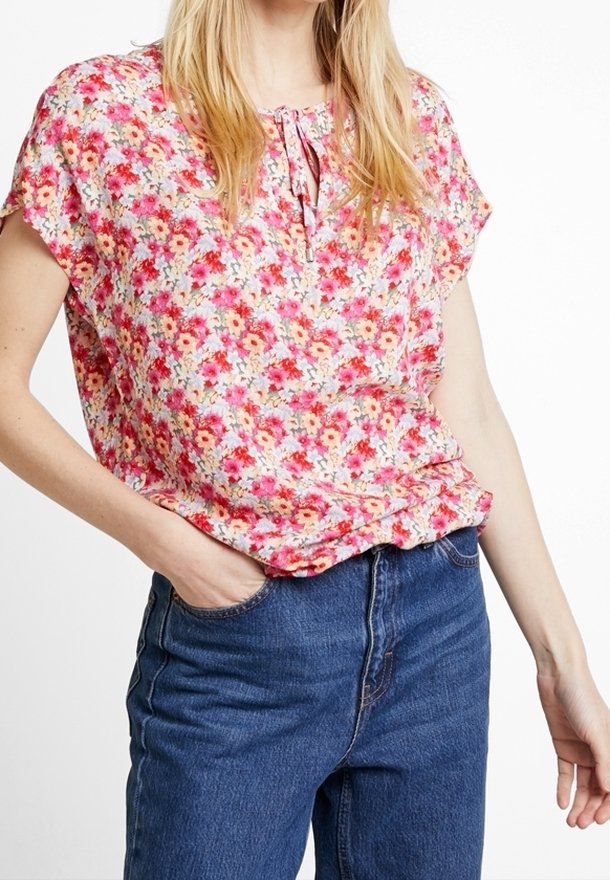 Blusa floral en rosa, naranja y amarillo con mangas cortas, cuello redondo y un detalle de lazo en el cuello, combinada con jeans de mezclilla.