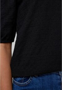 Svart texturerad topp med korta ärmar, prydd med ett mönster av små hål och sömnadsdetaljer, tillsammans med blå denimjeans.