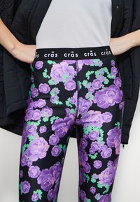 Leggings a vita alta con una base nera, caratterizzati da ampi motivi di rose viola bordati di verde e una cintura elastica nera con logo.