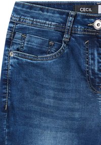 Jean en denim bleu foncé en gros plan montrant la poche avant, la petite poche à monnaie, le bouton, la boucle de ceinture et l’étiquette de marque "CECIL" sur la ceinture.