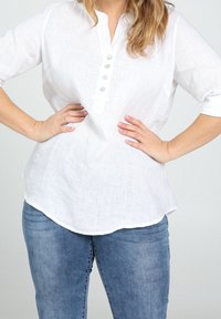 Chemise en lin blanc avec devant boutonné, manches trois-quarts, et ourlet arrondi. Associée à un jean denim bleu.