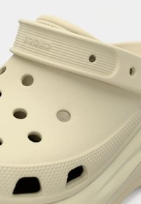 Gros plan d'un sabot Crocs beige montrant des trous de ventilation et une bride ajustable avec des rainures texturées.