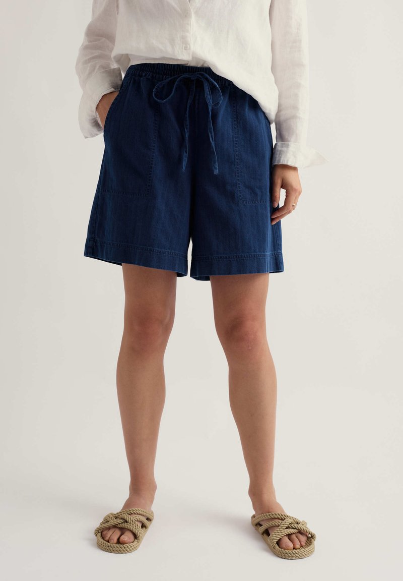 Frau trägt weißes Hemd mit Knöpfen, marineblaue Shorts mit Kordelzug und Vordertaschen sowie beige Sandalen aus Seil, steht vor weißem Hintergrund.