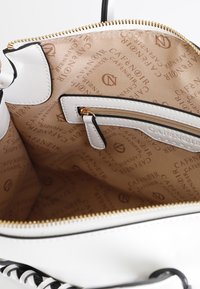 Hvid taske indvendigt med en beige stofforing med gentaget logomønster, en lynlåslomme og guldhardwaredetaljer.