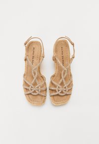 Beige sandaler med flat såle, med kryssende metallstropper og vevet kant. Spenne ved ankelen forbedrer passformen.