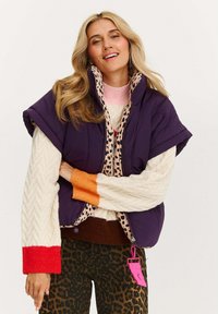 Fioletowa puchowa kamizelka z krótkimi rękawami i leopardzim wzorem w podszewce, noszona na jasnej sweterku z warkoczowym splotem. Brązowe spodnie w leopardzie wzór z różową metką.