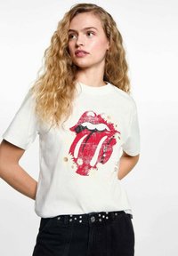 Biela bavlnená tričko s poškodeným červeným logom Rolling Stones a čiernymi akcentmi, doplnené o kvetinové a zlaté dekoratívne prvky.