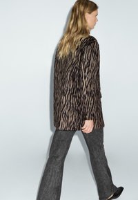 Dierenprint jas met een gemêleerd bruin en zwart patroon, gecombineerd met wijd uitlopende donkere jeans en zwarte schoenen. Model staat in zijaanzicht.