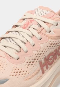 Αθλητικό παπούτσι σε απαλό ροζ χρώμα με πλέγμα στην επάνω πλευρά, μπεζ κορδόνια και υφή στη σόλα. Φέρει σήμανση HOKA και διακοσμητικά στοιχεία σε εναλλασσόμενη απόχρωση.