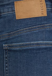 Dunkelblaue Denim-Jeans mit kontrastierendem braunem Nähten und einem rechteckigen, hellbraunen Lederetikett, das auf der Rückseite aufgenäht ist. Fünf-Taschen-Design.