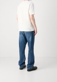 Bílé tričko s krátkým rukávem v kombinaci s modrými džíny, které mají volný střih a rovné nohavice. Celkový vzhled doplňují černé boty.