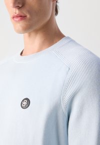 Light blue knit sweater featuring a ribbed crew neckline and side accents, with a black and silver logo patch on the chest.