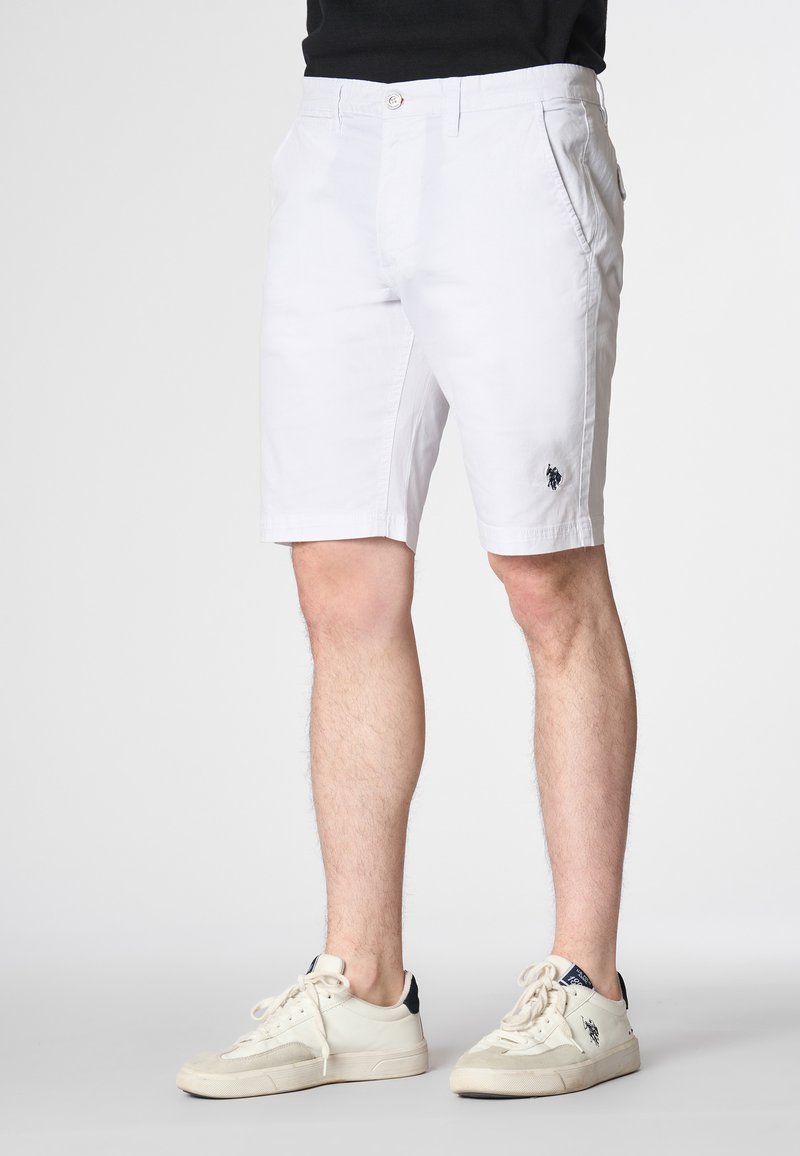 Pantaloni corti bianchi in cotone con vestibilità dritta, tasche frontali e logo ricamato sulla gamba sinistra. Indossati con sneakers beige e una maglietta nera.
