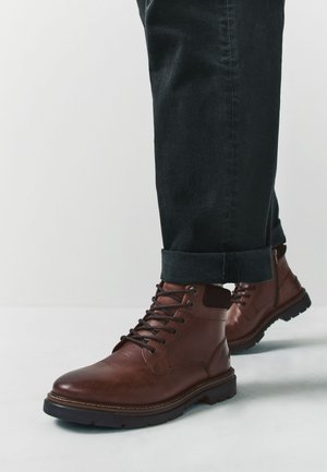 Braune Lederstiefel mit Schnürsenkeln, ausgestattet mit einer runden Zehenpartie und einer strukturierten Oberfläche. Schwarze Denim-Hose mit umgeschlagenem Saum zeigt das Design des Stiefels.