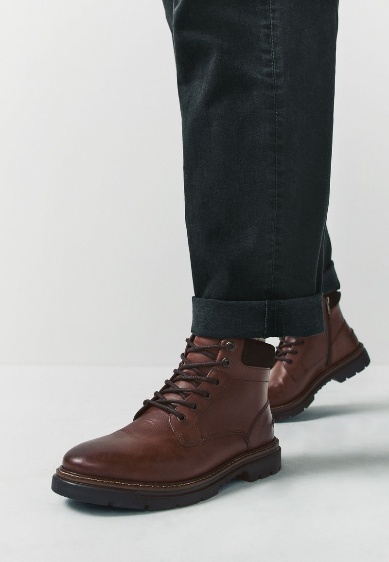 Braune Lederstiefel mit Schnürsenkeln, ausgestattet mit einer runden Zehenpartie und einer strukturierten Oberfläche. Schwarze Denim-Hose mit umgeschlagenem Saum zeigt das Design des Stiefels.