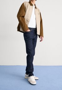 Hombre con chaqueta de bloques de color marrón y beige, camiseta blanca, vaqueros azul oscuro y zapatillas beige, de pie sobre un suelo alfombrado azul.