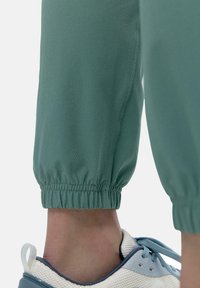 Grønne, letvægts joggere med en tekstureret finish, der har elastiske manchetter. Anklerne er parret med en hvid og blå træningssko.