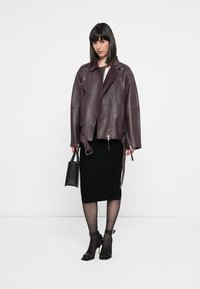 Veste de moto en cuir bordeaux avec des détails zippés, portée sur une jupe noire texturée. Les accessoires incluent un petit sac à main noir et des talons.