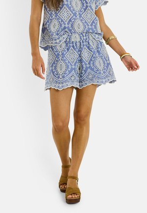Mujer que lleva pantalones cortos y top a juego con patrón azul y bordes ondulados, complementados con pulseras doradas y sandalias marrones.
