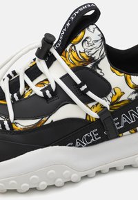 Baskets noires et blanches avec un motif floral, dotées d'une semelle en caoutchouc texturée, de lacets élastiques et d'un logo bien visible sur le côté.