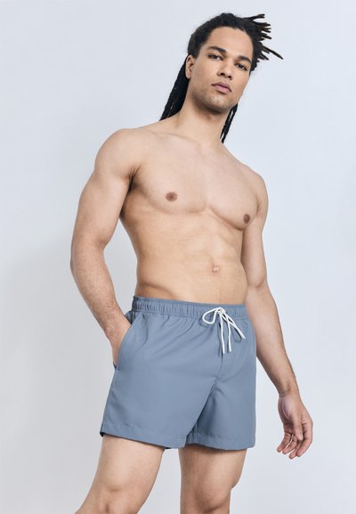Shirtless man with long hair in blue drawstring shorts stands with one hand in pocket against plain background.